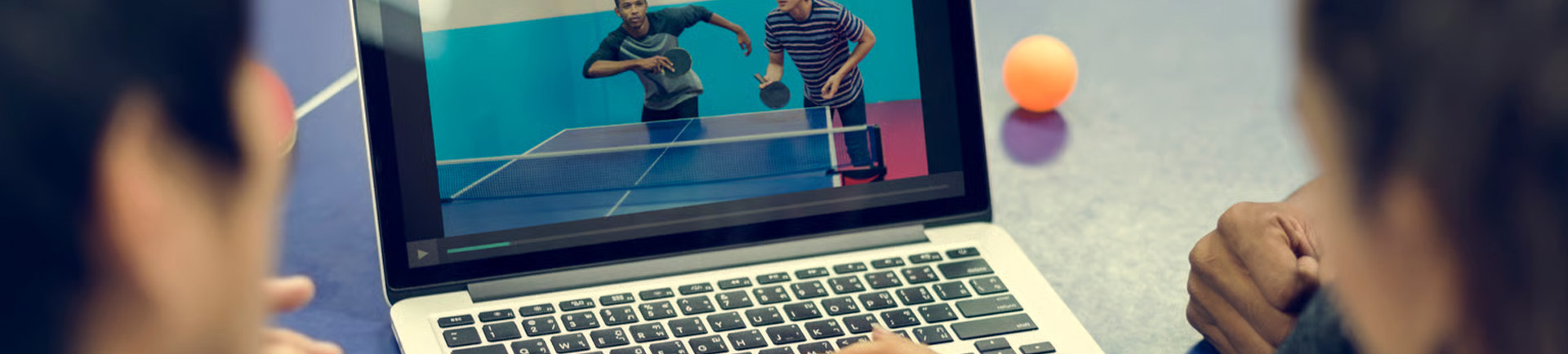 Duas cabeças vistas de trás, à frente de um computador com uma imagem de dois jovens a jogar ping pong.