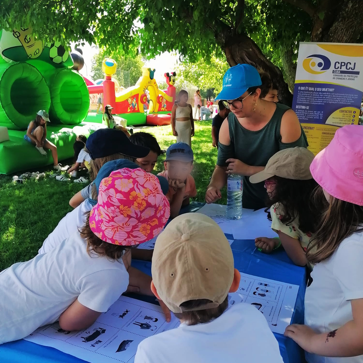 Técnica do IPDJ centrada na fotografia, numa mesa rodeada de crianças e folhas de papel à frente de cada uma, focadas na atividade. A técnica explica a atividade. No fundo pode-se ver-se o cenario de um jardim com insufláveis.