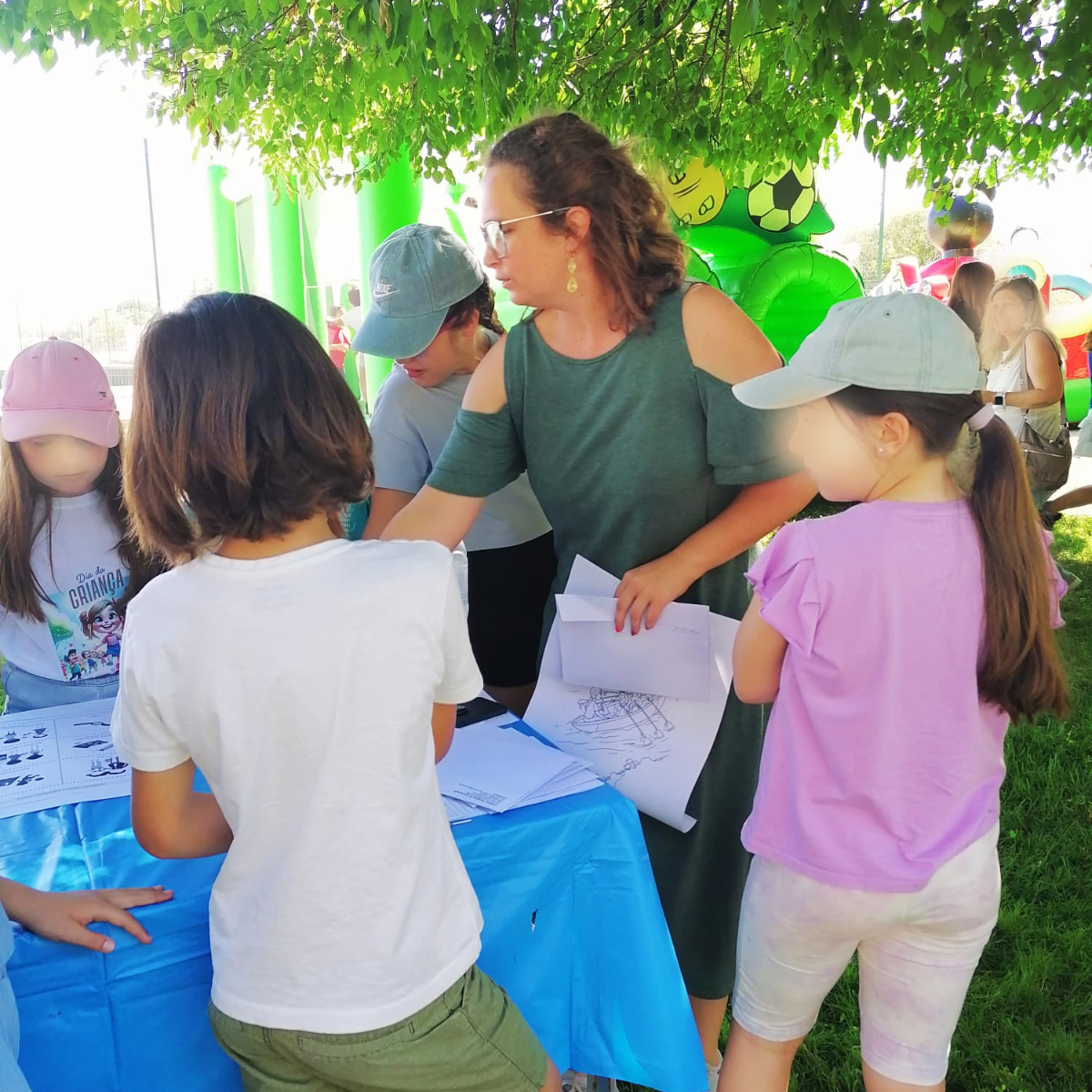 A técnica encontra-se com folhas nas mãos, a explicar a atividade às crianças junto da mesa de atividades. Cenário de fundo no jardim.