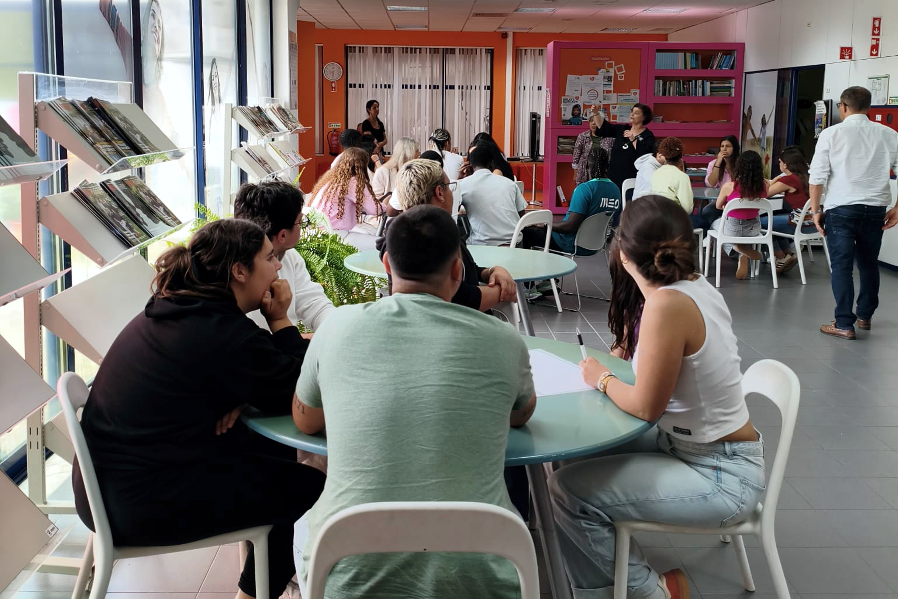 Grupos de jovens sentados em mesas redondas a ouvir as instruções da técnica relativamente à atividade que vão desenvolver.