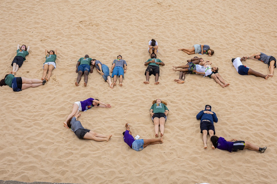 Foto de jovens na praia a desenhar com o corpo a letra CJL