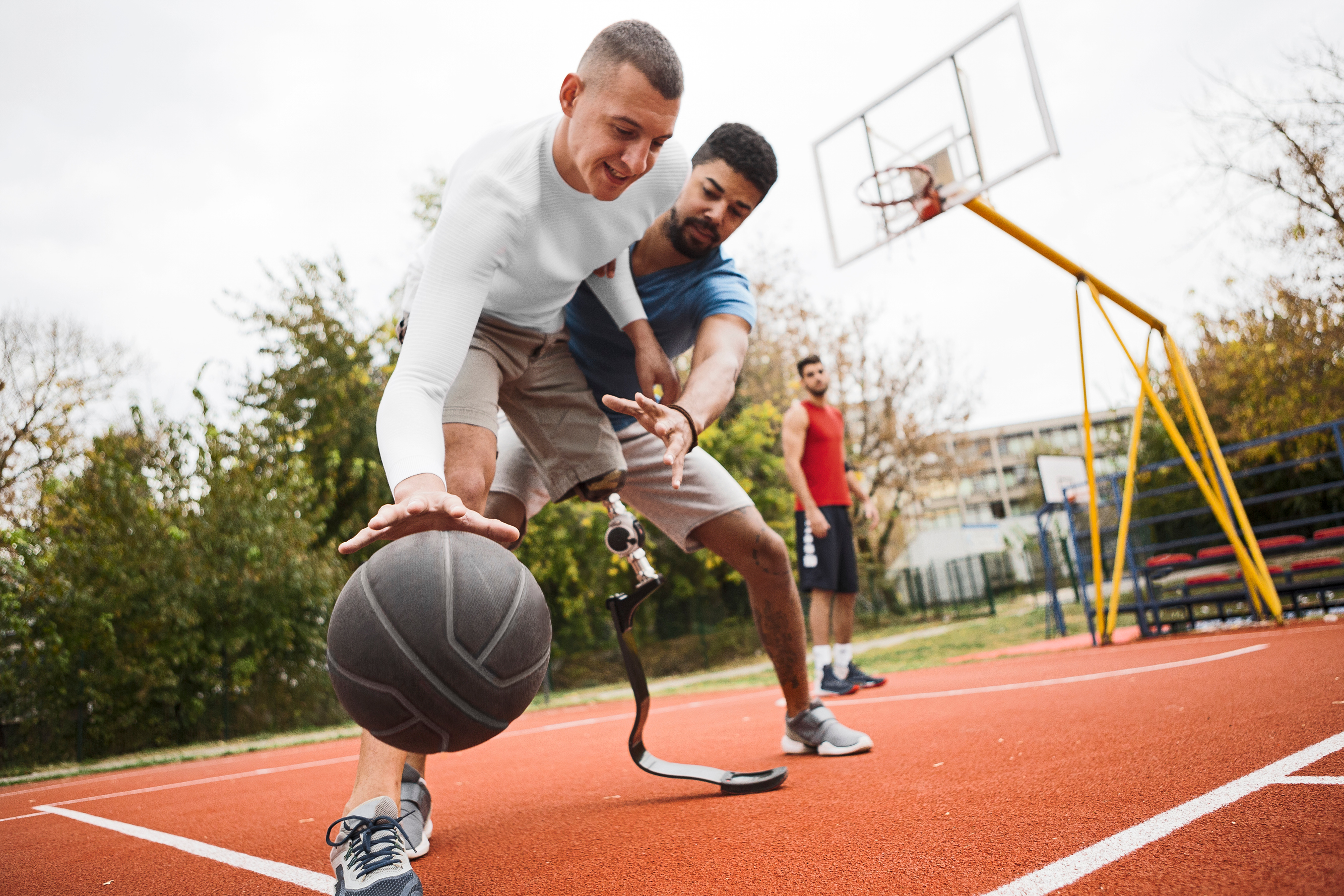 Três desportistas masculinos a jogar basquetebol, um deles tem deficiência motora num dos membros inferiores.