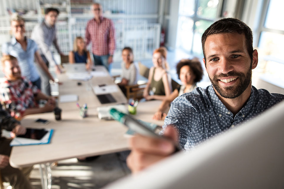 Um jovem a escrever num flipshart, com um grupo de jovens sentados atrás.