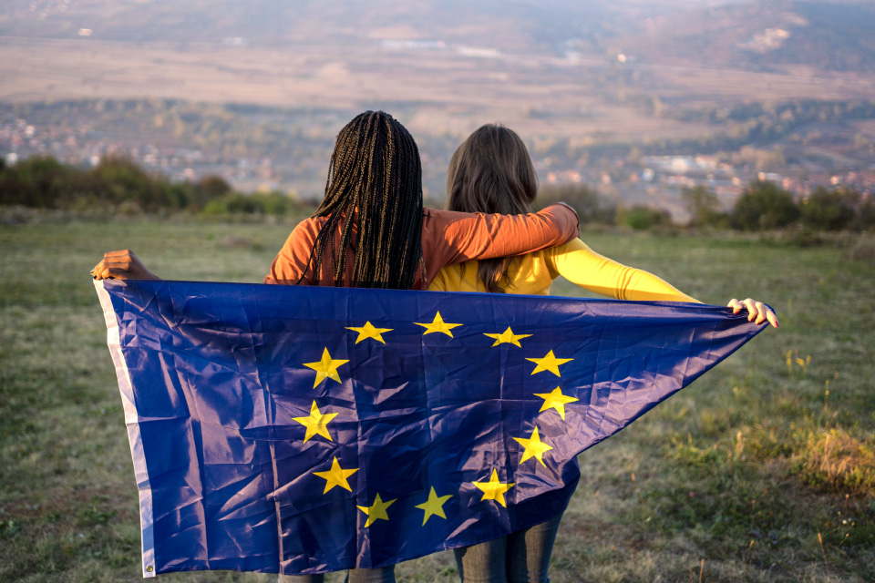 Duas jovens de costas com a bandeira da UE, numa paisagem verde. 