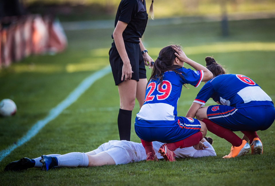 Três raparigas - uma mulher árbitro -, e duas jogadoras num campo relvado, uma delas deitada com sinais de dor.