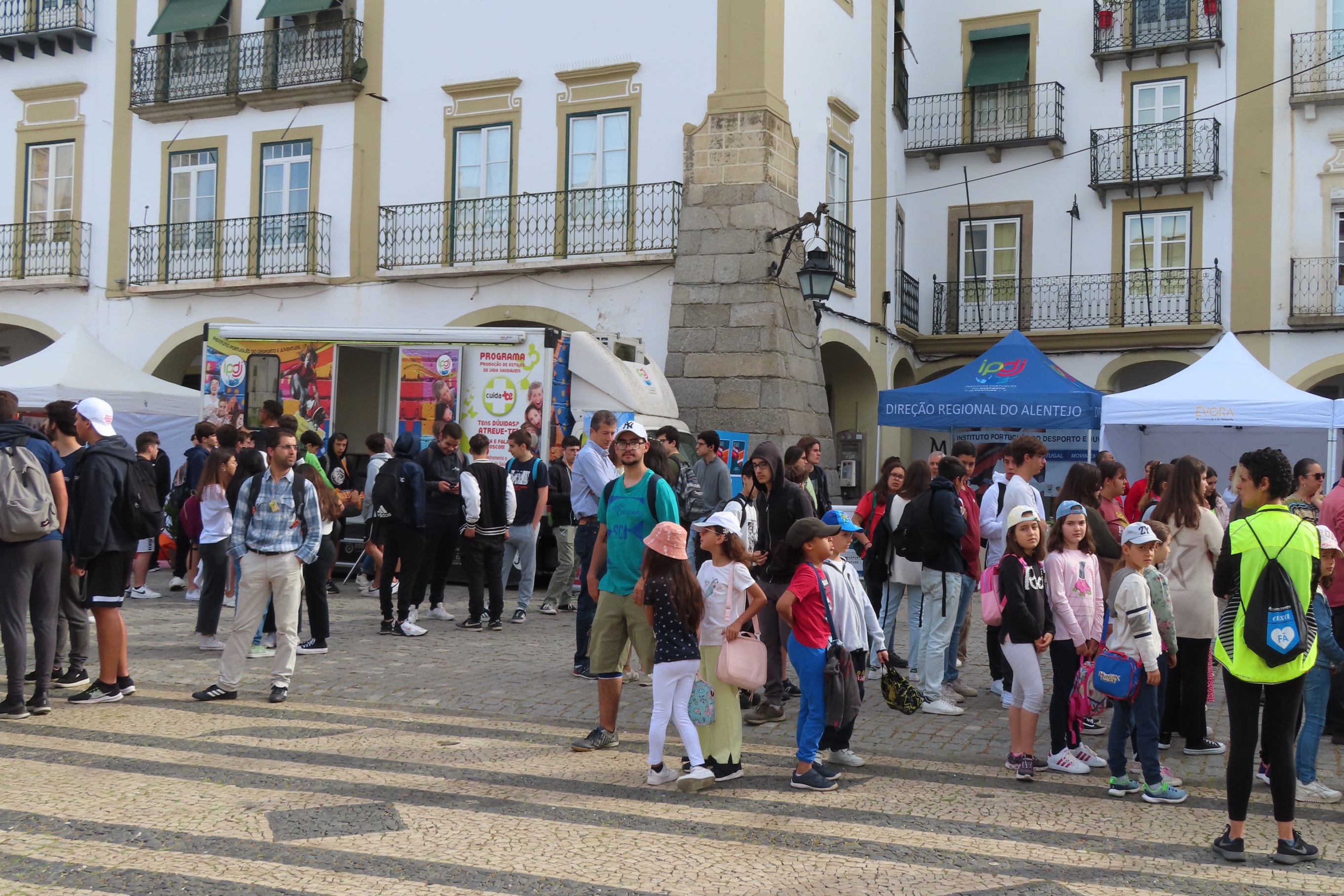 Várias pessoas na Praça do Giraldo, junto à Unidade Móvel Cuida-te +, e junto ao toldo IPDJ.