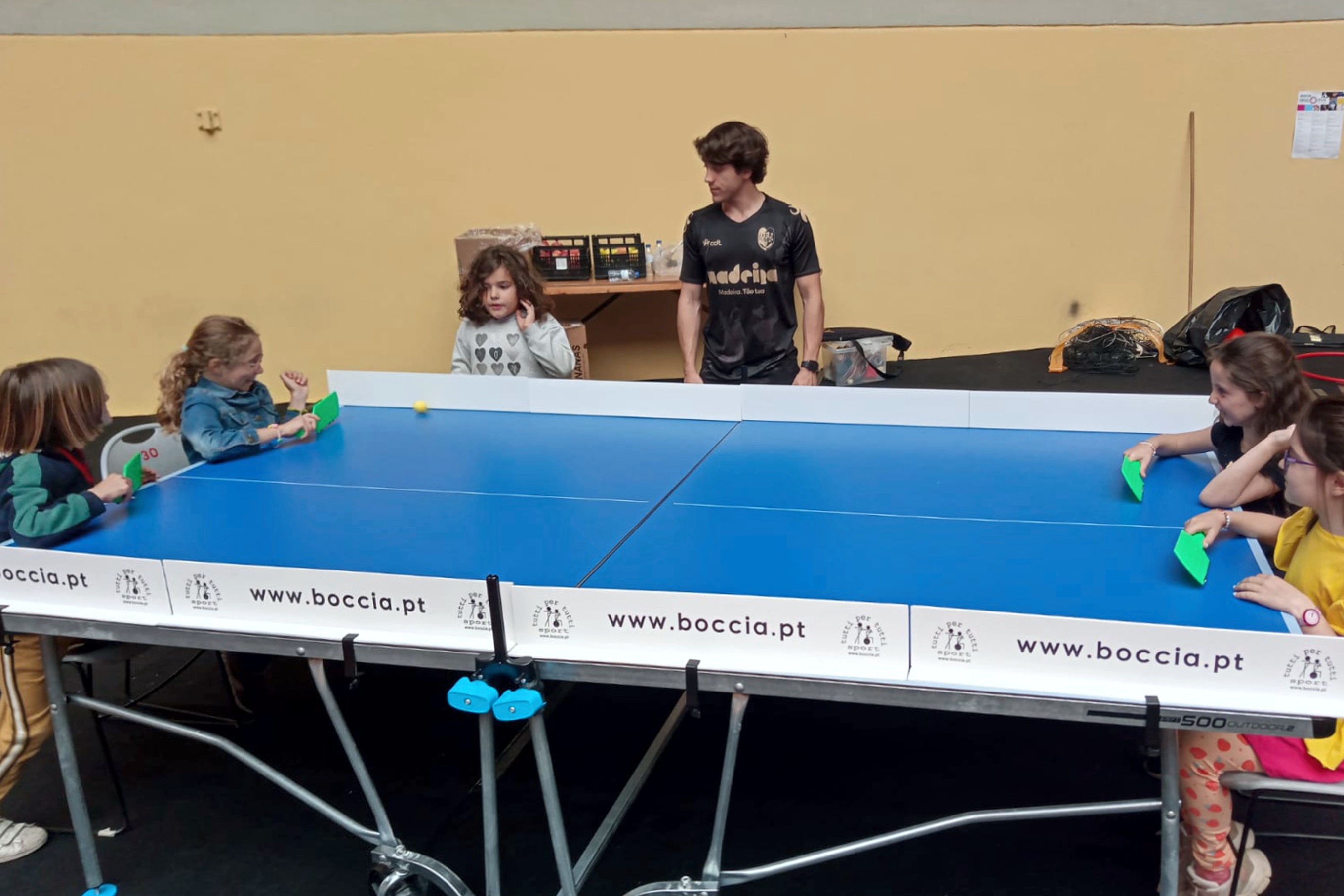 Várias crianças e um jovem voluntário, na Arena D' Évora, a jogarem ténis de mesa.