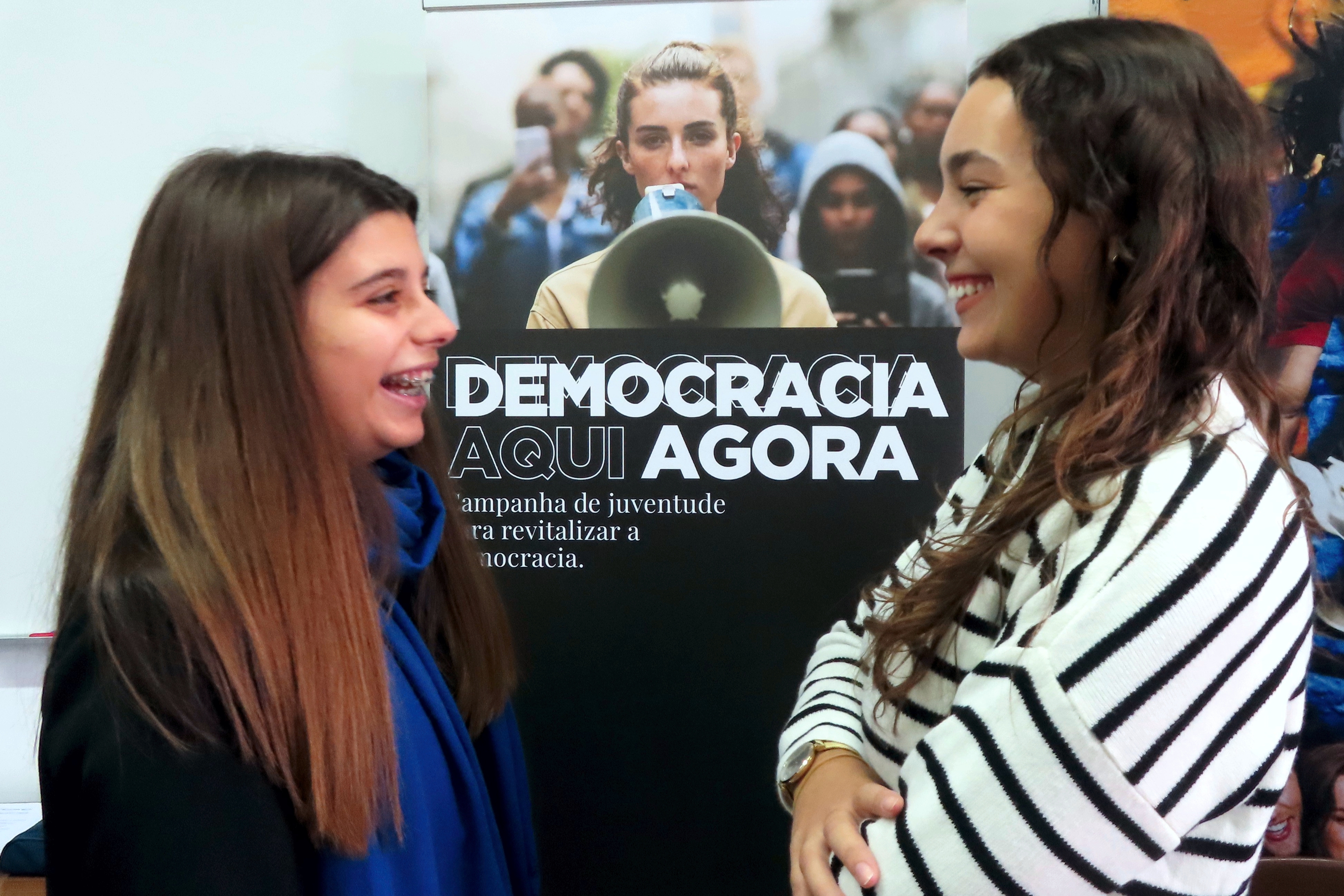 As voluntárias Ana Rita Coelho e Mariana Costa, tendo como painel de fundo o Roll-Up da Campanha «Democracia Aqui. Democracia Agora».