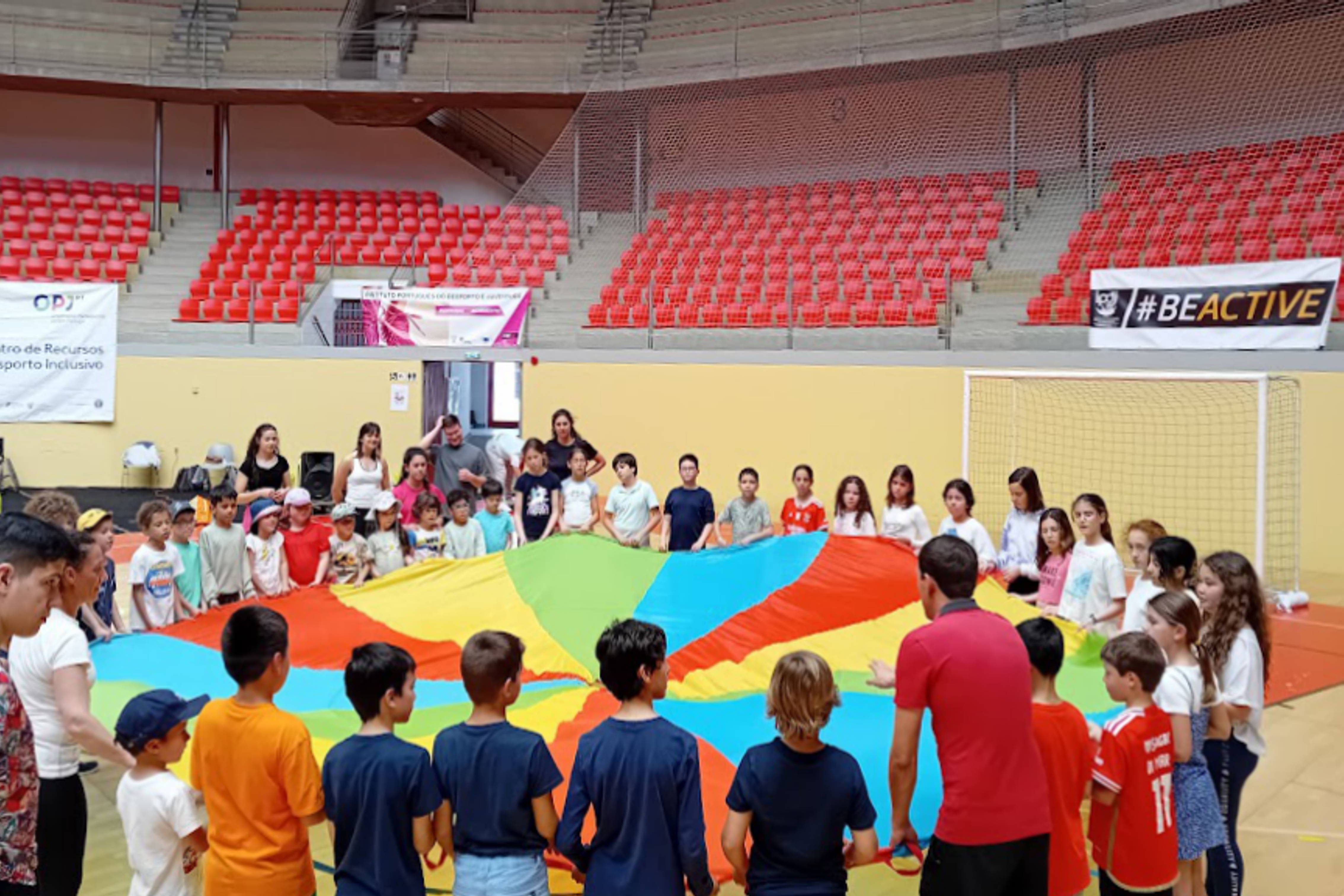 Grupo de alunos/as e professores/as, na Arena D'Évora, a participarem numa dinâmica de atividade física, com um páraquedas recreativo.