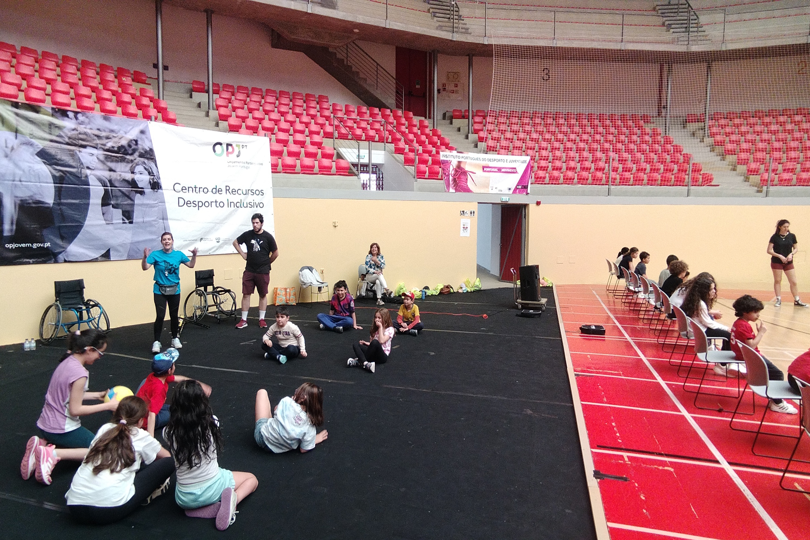 Grupo de crianças na Arena D'Évora, a participarem numa dinâmica de atividade física, com uma bola, dinamizada pela técnica do IPDJ de Évora, Andreia Peixe com a colaboração de um aluno da Universidade de Évora.