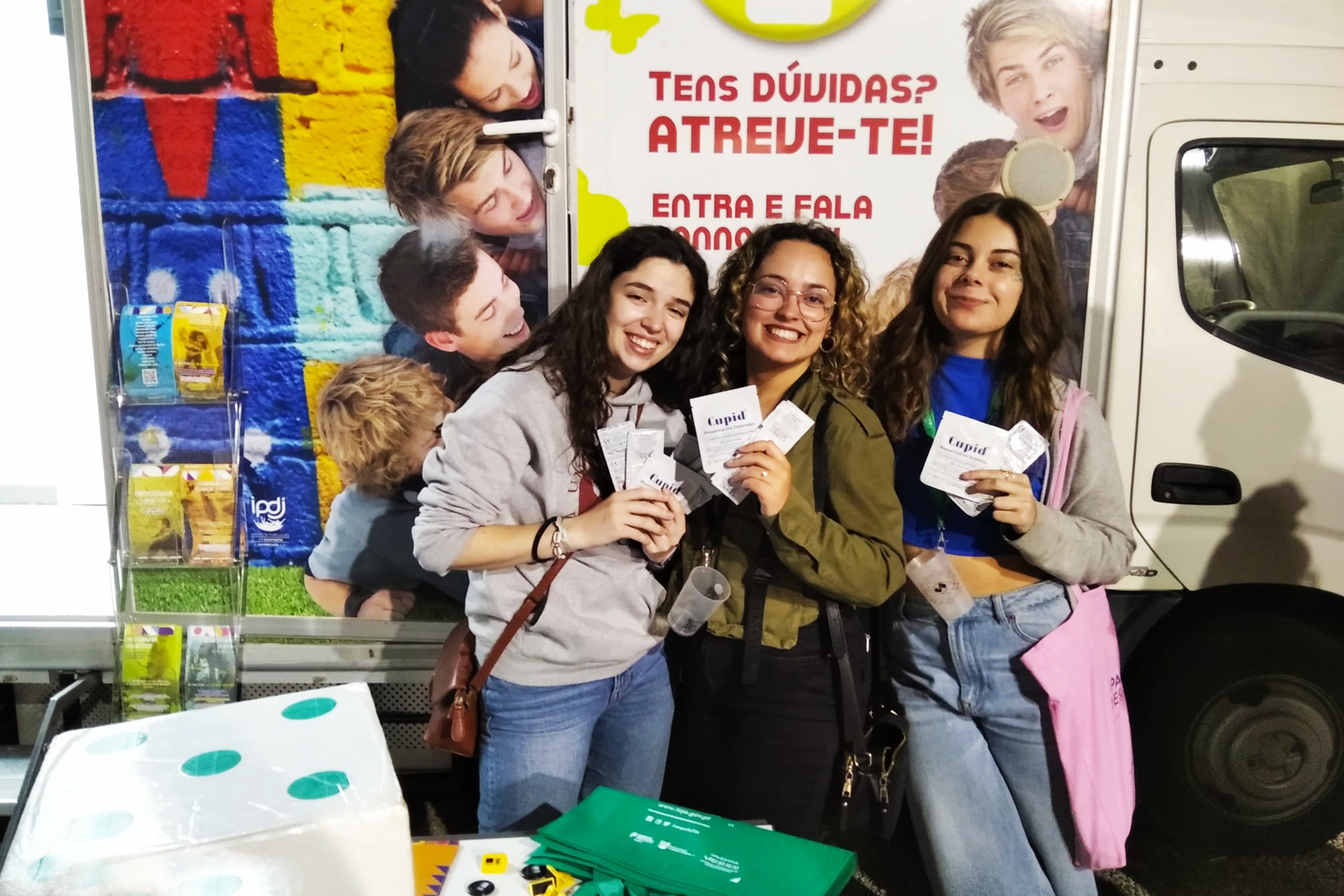 Três jovens (raparigas) junto da Unidade Móvel Cuida-te+, a mostrarem cada uma delas os preservativos masculinos e femininos, nas mãos.
